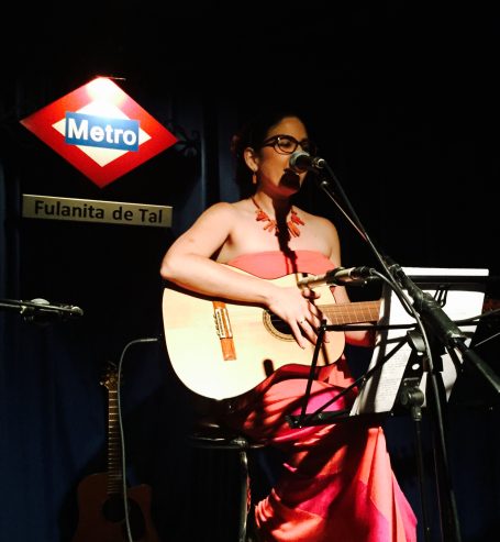 Mujer con guitarra en un escenario, vestida de rosa, cantando en un ambiente nocturno.