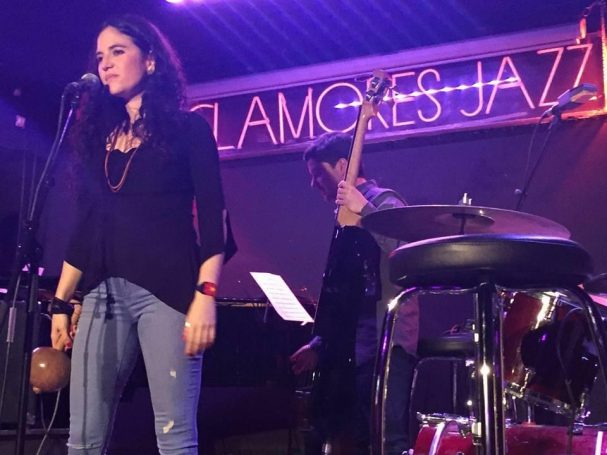 Mujer canta en un escenario de jazz, con fondo iluminado y un músico detrás.