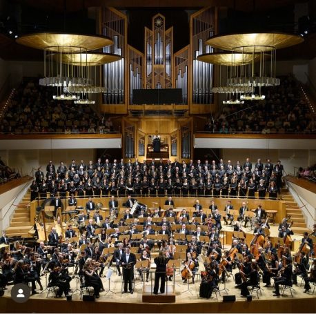 Orquesta en un auditorio, con músicos en el escenario y un director dirigiendo.