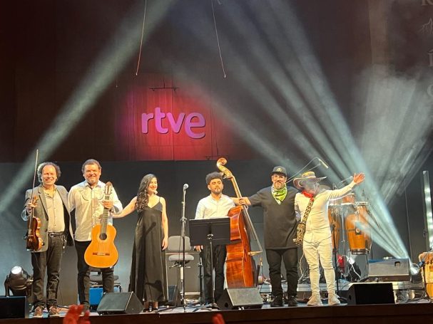 Grupo musical en el escenario, saludando al público con instrumentos y micrófonos.