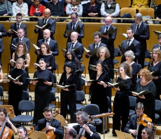 Coros de pie en un escenario, cantando con partituras, rodeados de músicos.