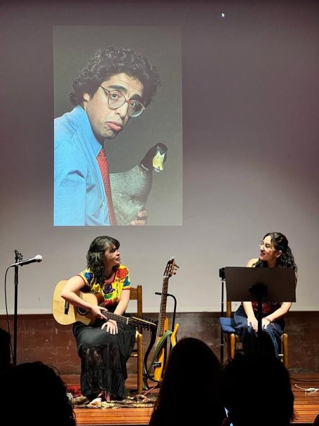 Una mujer con guitarra y otra sentada, con una imagen de fondo de un hombre con gafas y un pingüino.